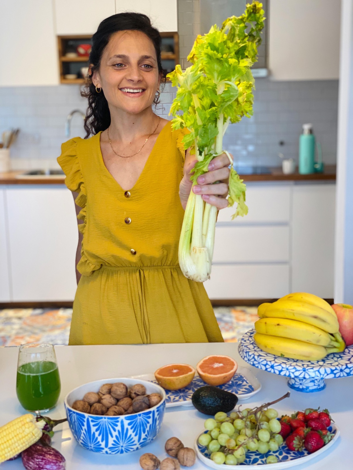 Juli sonriendo en su cocina con un apio fresco en la mano, frente a una mesa con frutas, nueces y jugo verde