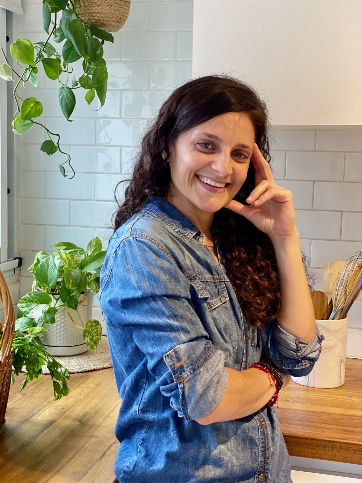 Retrato de Juli Carra sonriendo en su cocina, con camisa de jean y plantas de fondo